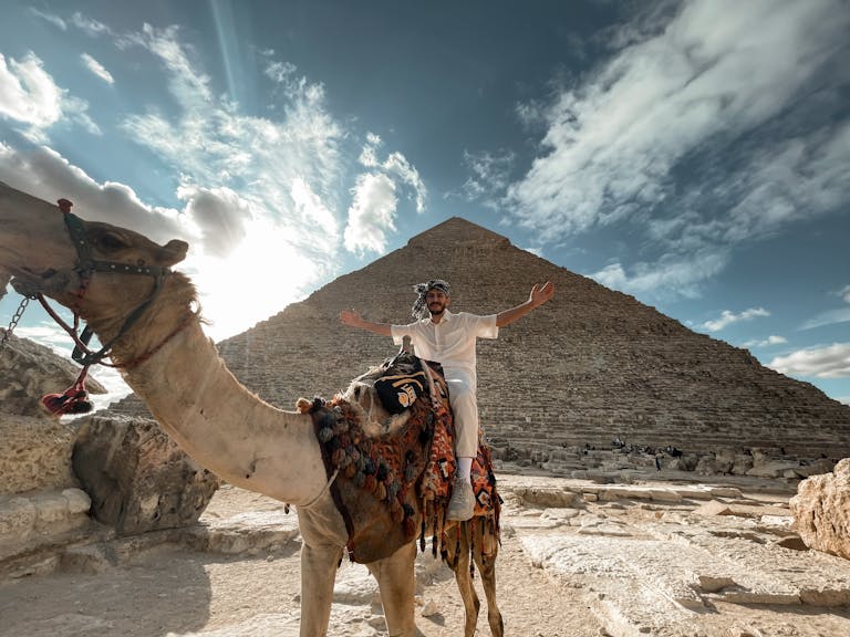 A man sitting on a camel in front of the Giza Pyramid during a stunning sunset, showcasing Egyptian landmarks.