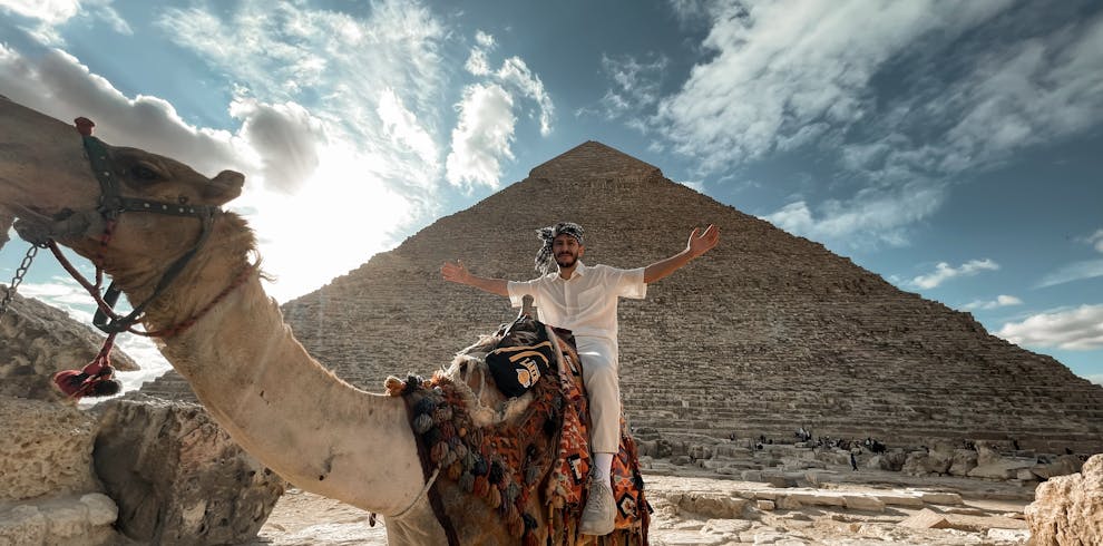 A man sitting on a camel in front of the Giza Pyramid during a stunning sunset, showcasing Egyptian landmarks.