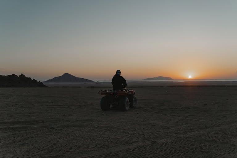 A person on an ATV rides through the desert at sunset, capturing the serene beauty of sand and distant mountains.