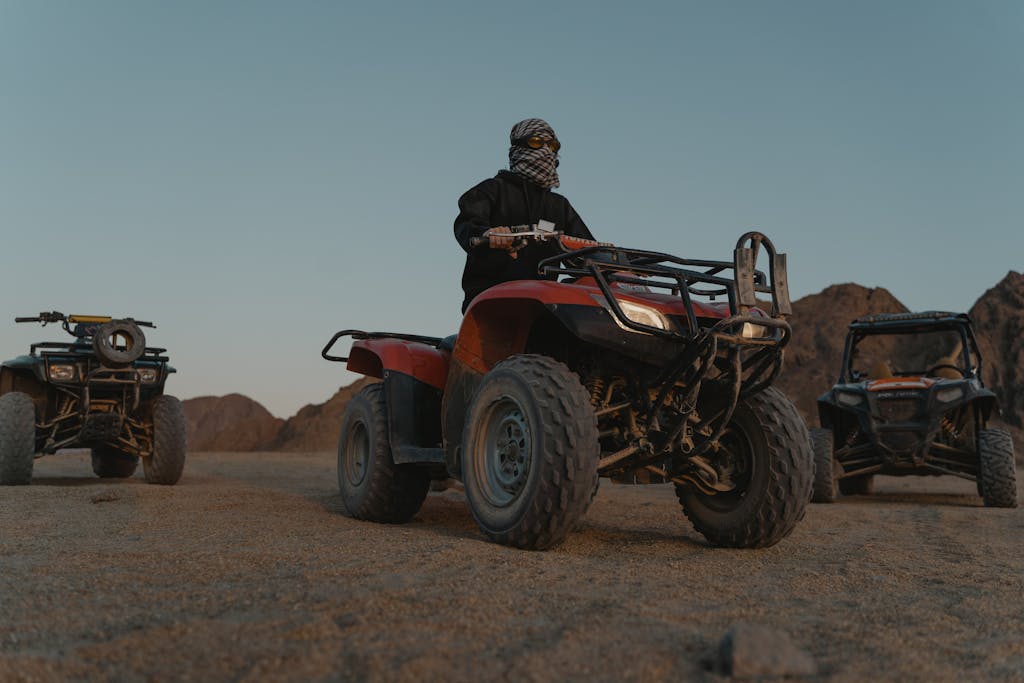 Riders on quad bikes explore a rugged desert terrain, with mountains in the background.